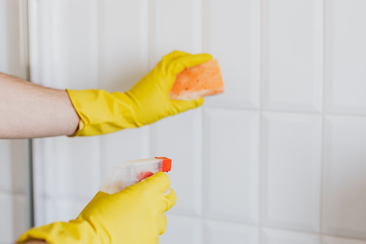 Crop unrecognizable woman in yellow rubber gloves cleaning white tiles with spray and soapy sponge